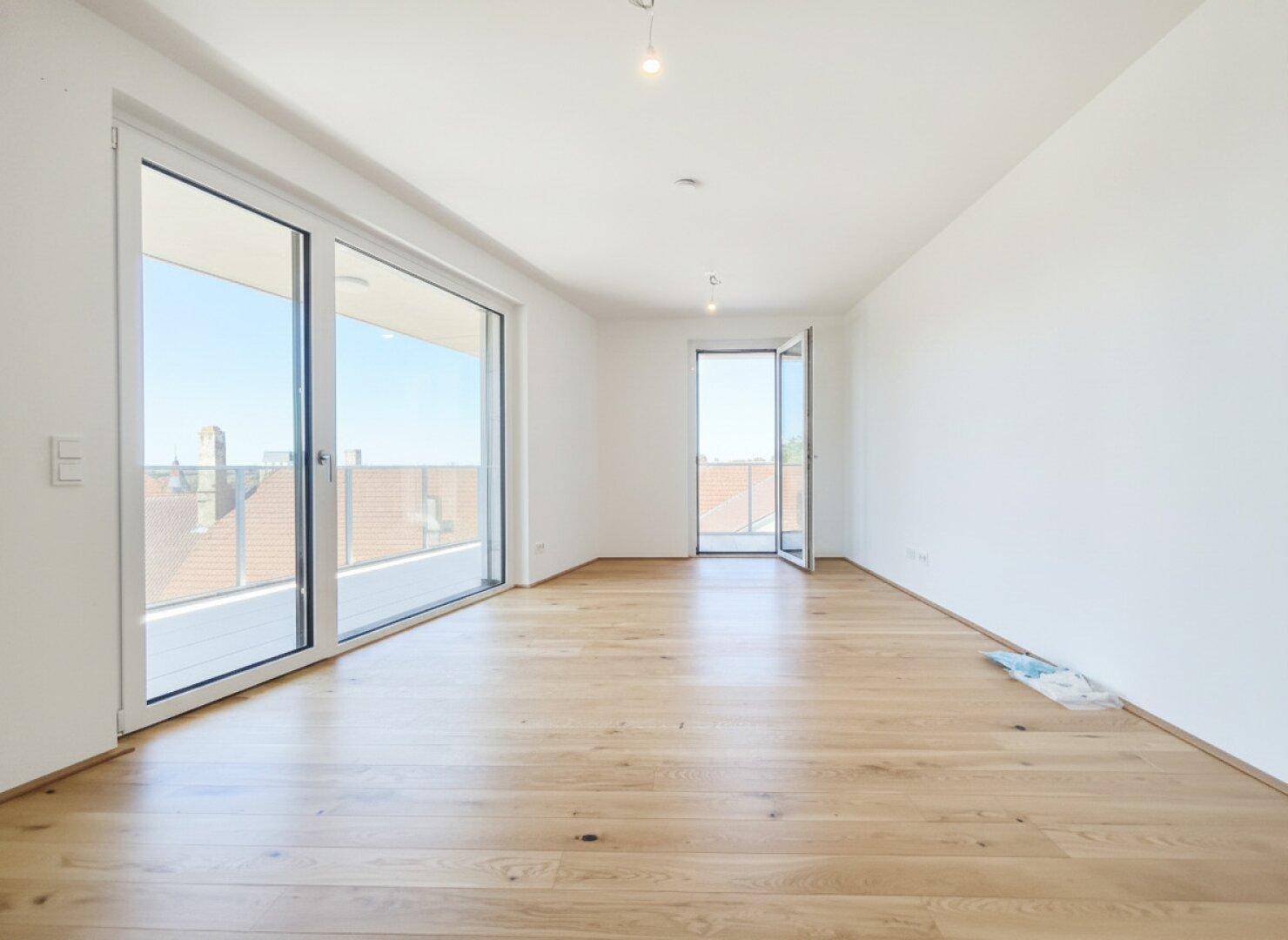 Lichtdurchfluteter Raum mit hellem Parkettboden und zwei großen Fenstertüren, die auf einen Balkon führen.