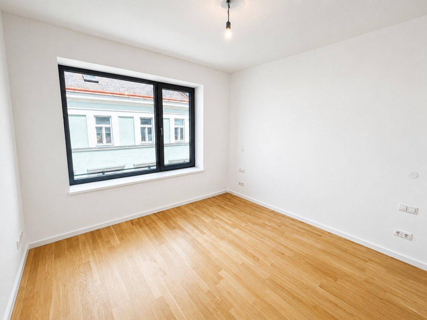 Freundliches Zimmer mit Holzboden und einem großen Fenster, das viel Tageslicht hereinlässt.