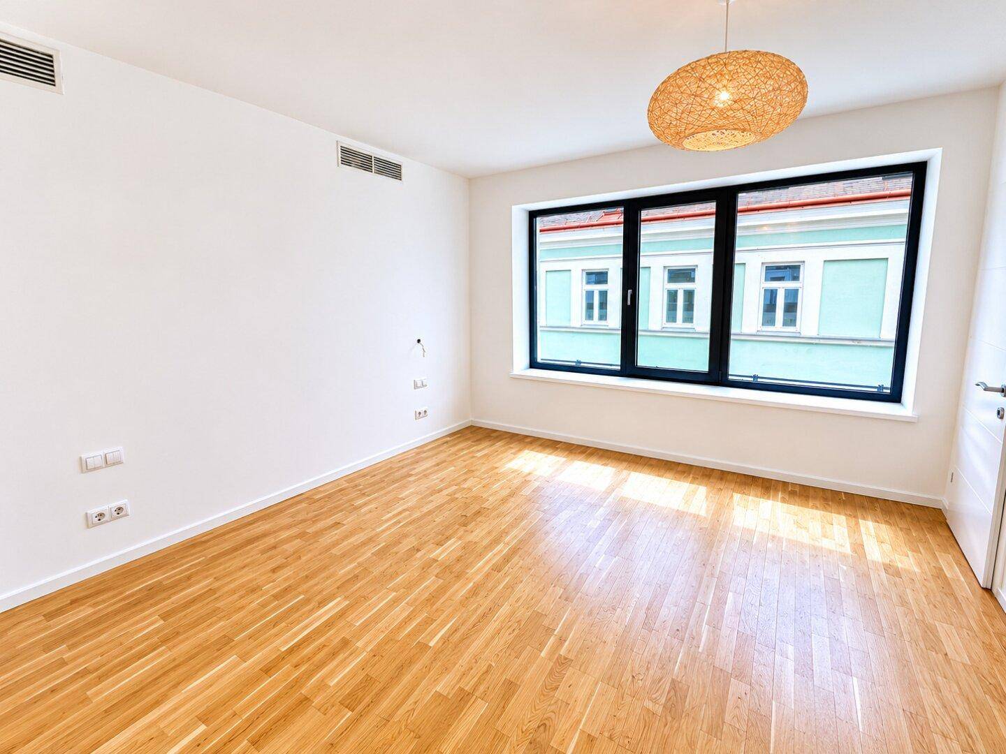 Leeres Zimmer mit Holzboden und großem Fenster mit Blick auf ein gegenüberliegendes Gebäude.