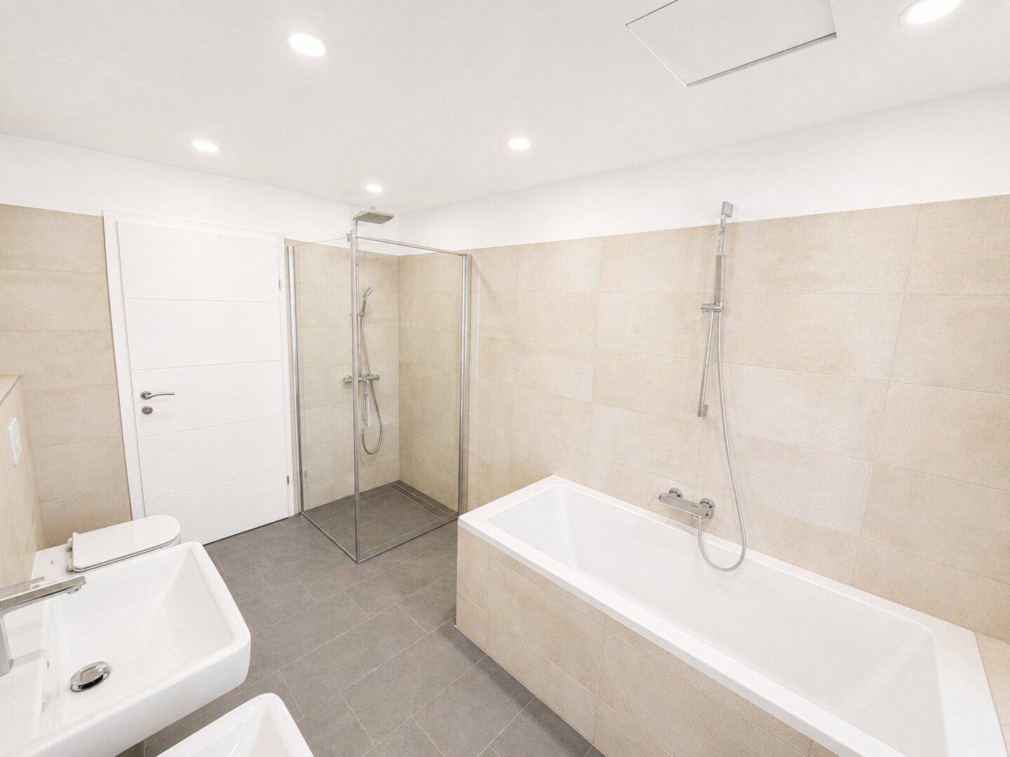 Helles Badezimmer mit beigen Wandfliesen, Badewanne, begehbarer Dusche und modernem WC.