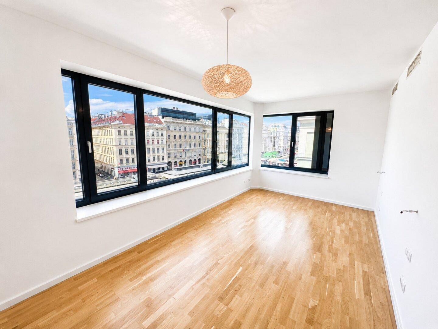 Helles Zimmer mit Holzboden, großen Fenstern und Blick auf die Stadt, unmöbliert.