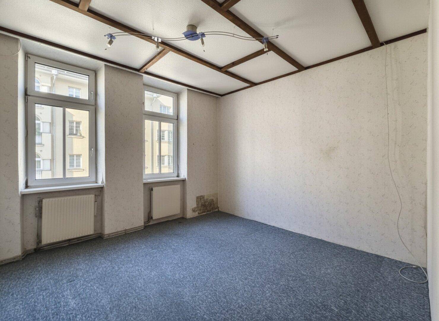 Unmöblierter Raum mit blauem Teppichboden, zwei Fenstern und sichtbaren Deckenbalken.