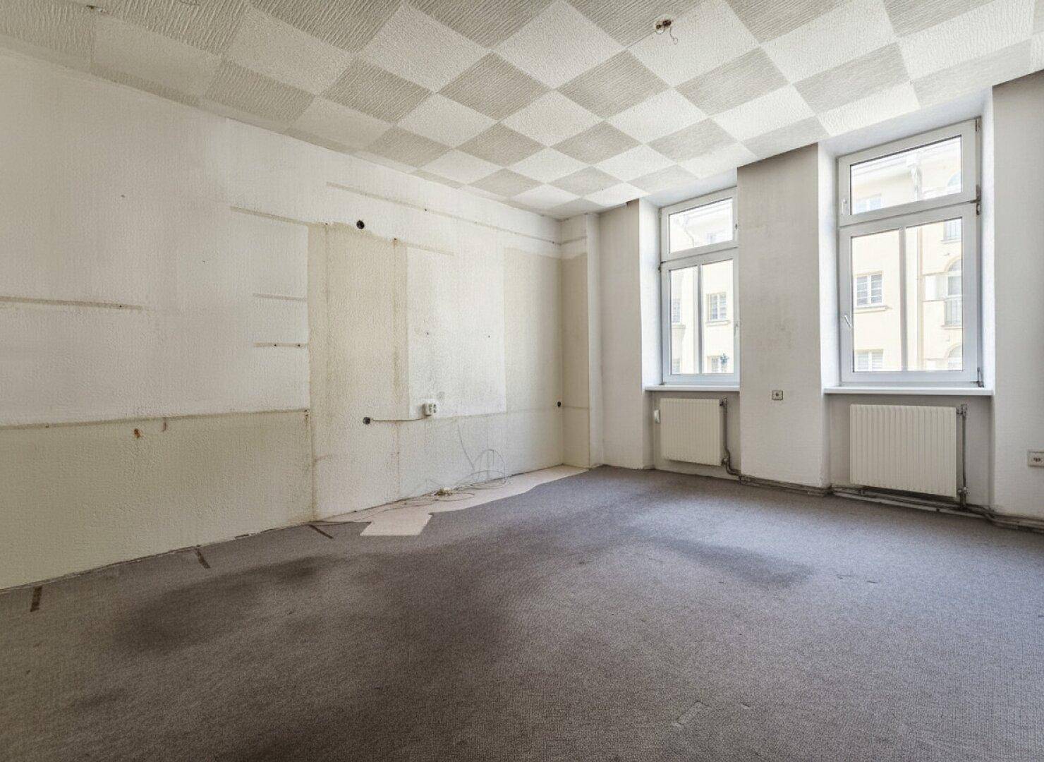 Unmöblierter Raum in Renovierungszustand mit Teppichboden und zwei Fenstern.