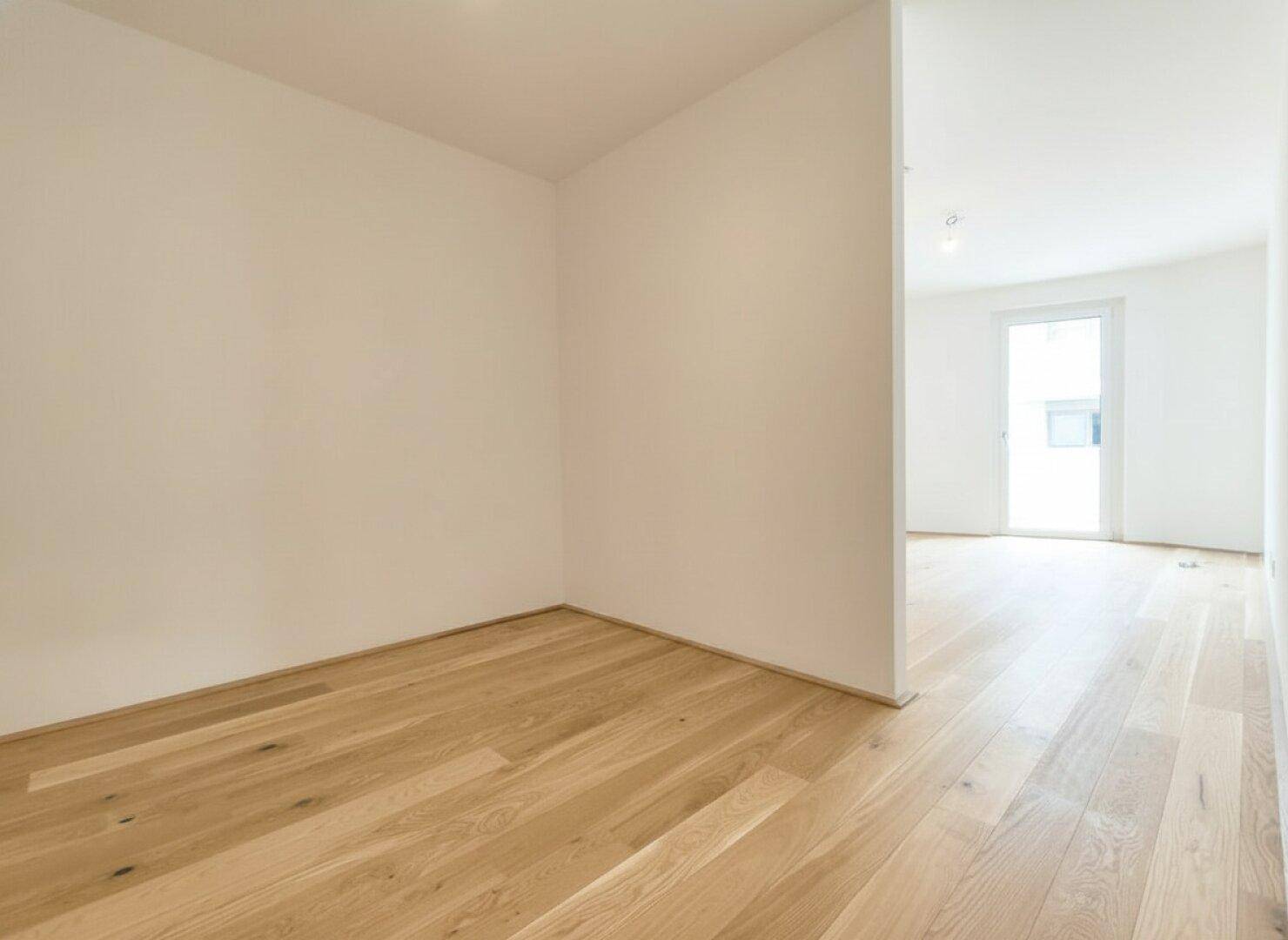 Heller Raum mit Holzparkett und weiß gestrichenen Wänden, der in einen weiteren Bereich übergeht.