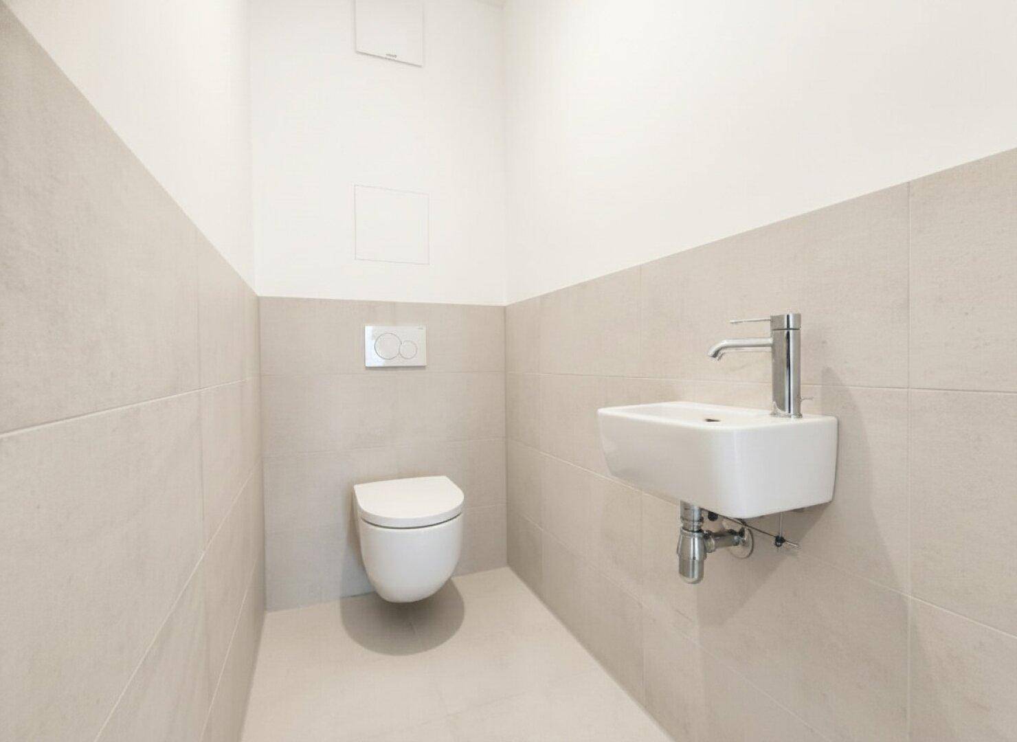 Modernes Gäste-WC mit hellgrauen Fliesen, wandhängender Toilette und kleinem Handwaschbecken.