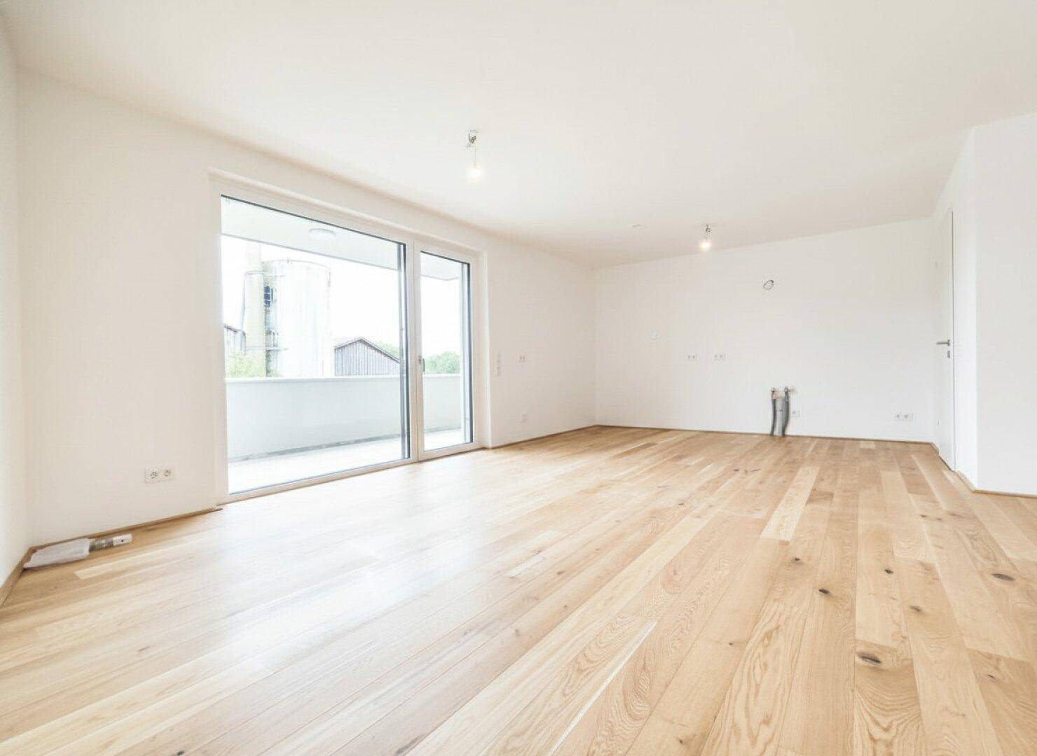 Lichtdurchflutetes Zimmer mit hellem Holzfußboden und großzügiger Schiebetür zum Balkon, bereit zum Einzug.