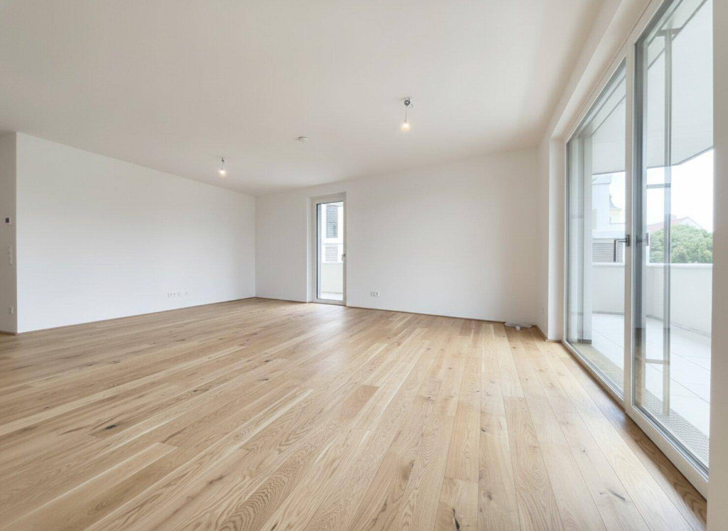 Helles, leeres Zimmer mit hellem Holzparkett und Zugang zu einem großen Balkon durch eine Glasschiebetür.