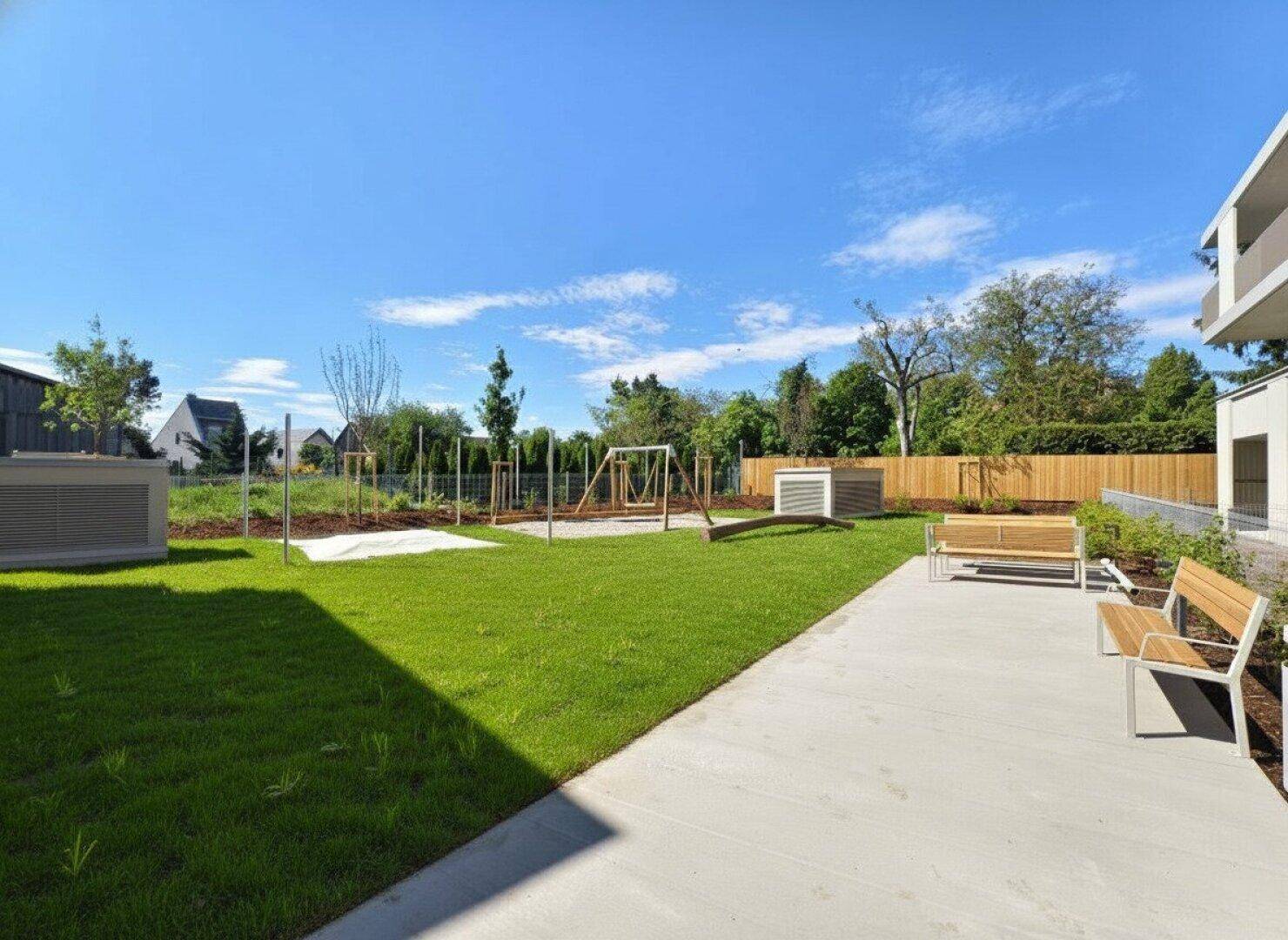 Gemeinschaftlicher Außenbereich mit gepflegtem Rasen, Sitzbänken und einem Spielplatz im Hintergrund.