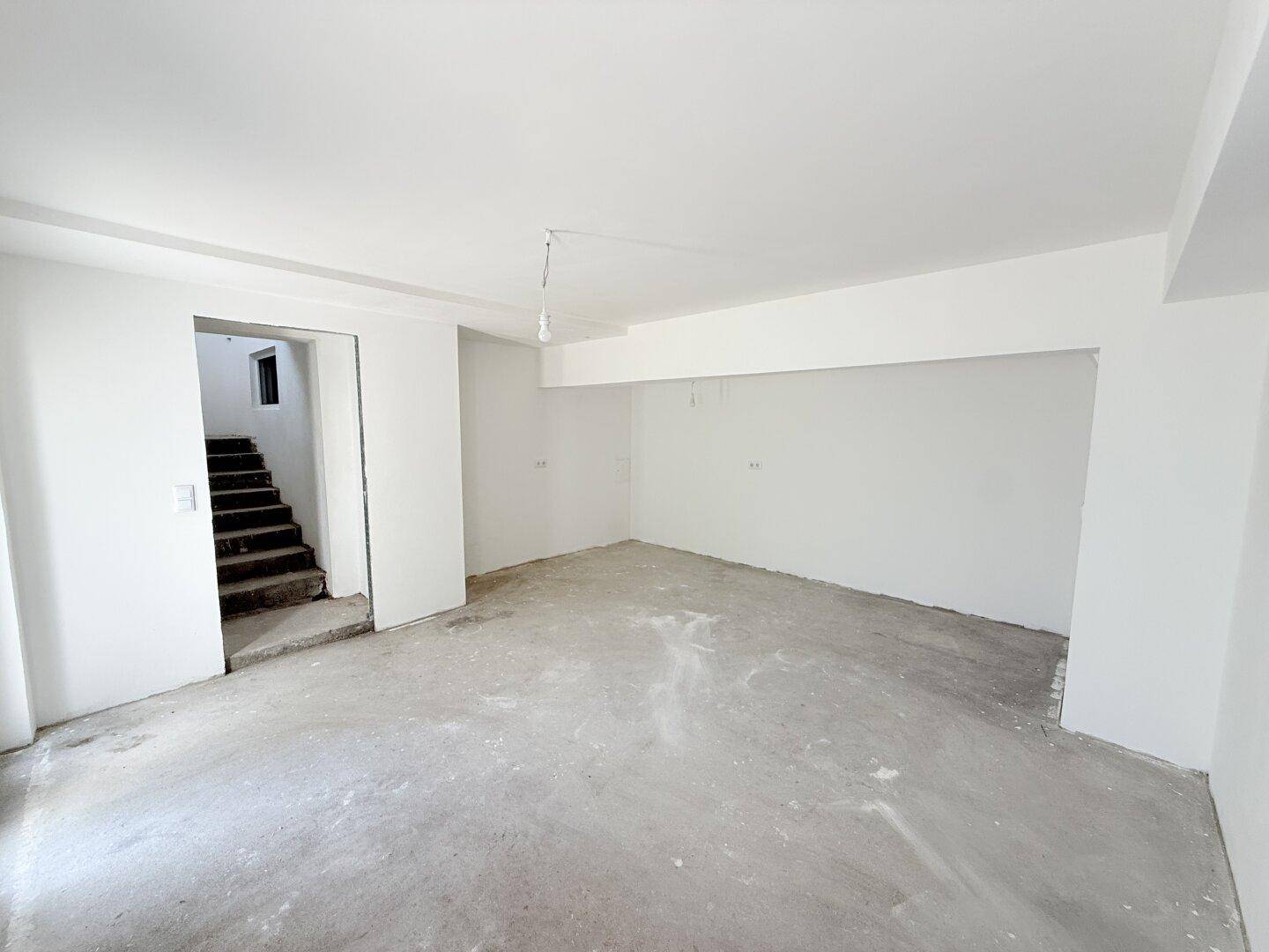 Geräumiger, unfertiger Raum mit weiß gestrichenen Wänden und Zugang zu einer Betontreppe.