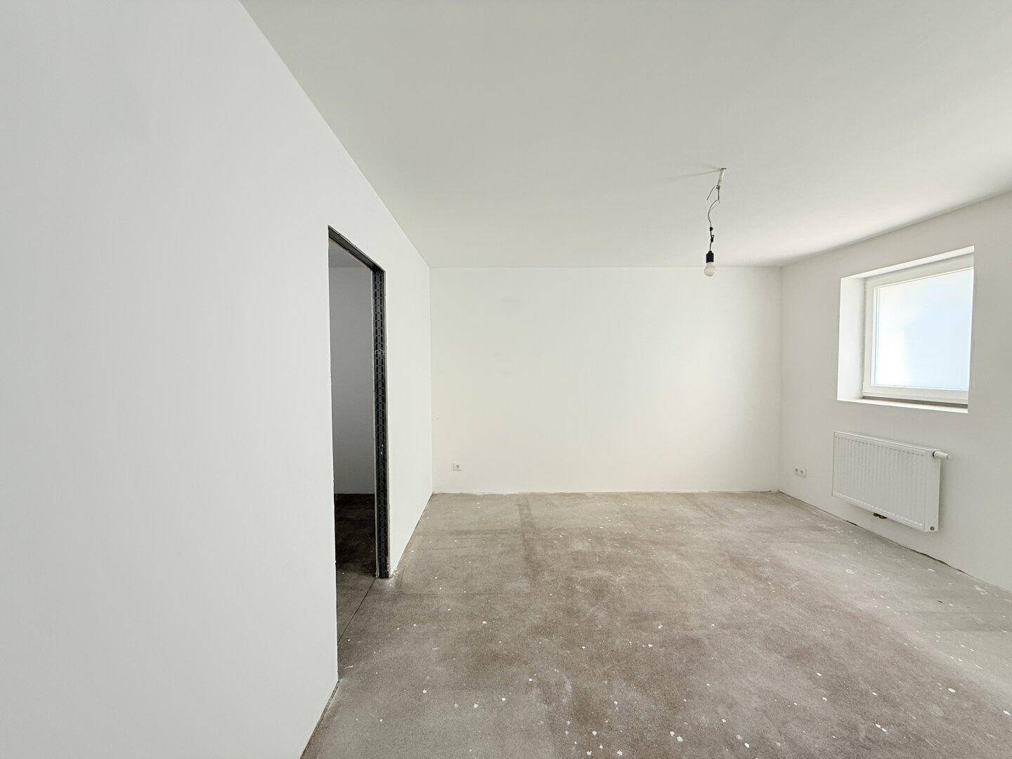 Geräumiger, unfertiger Raum mit einem Fenster und Heizkörper, bereit für den Innenausbau.