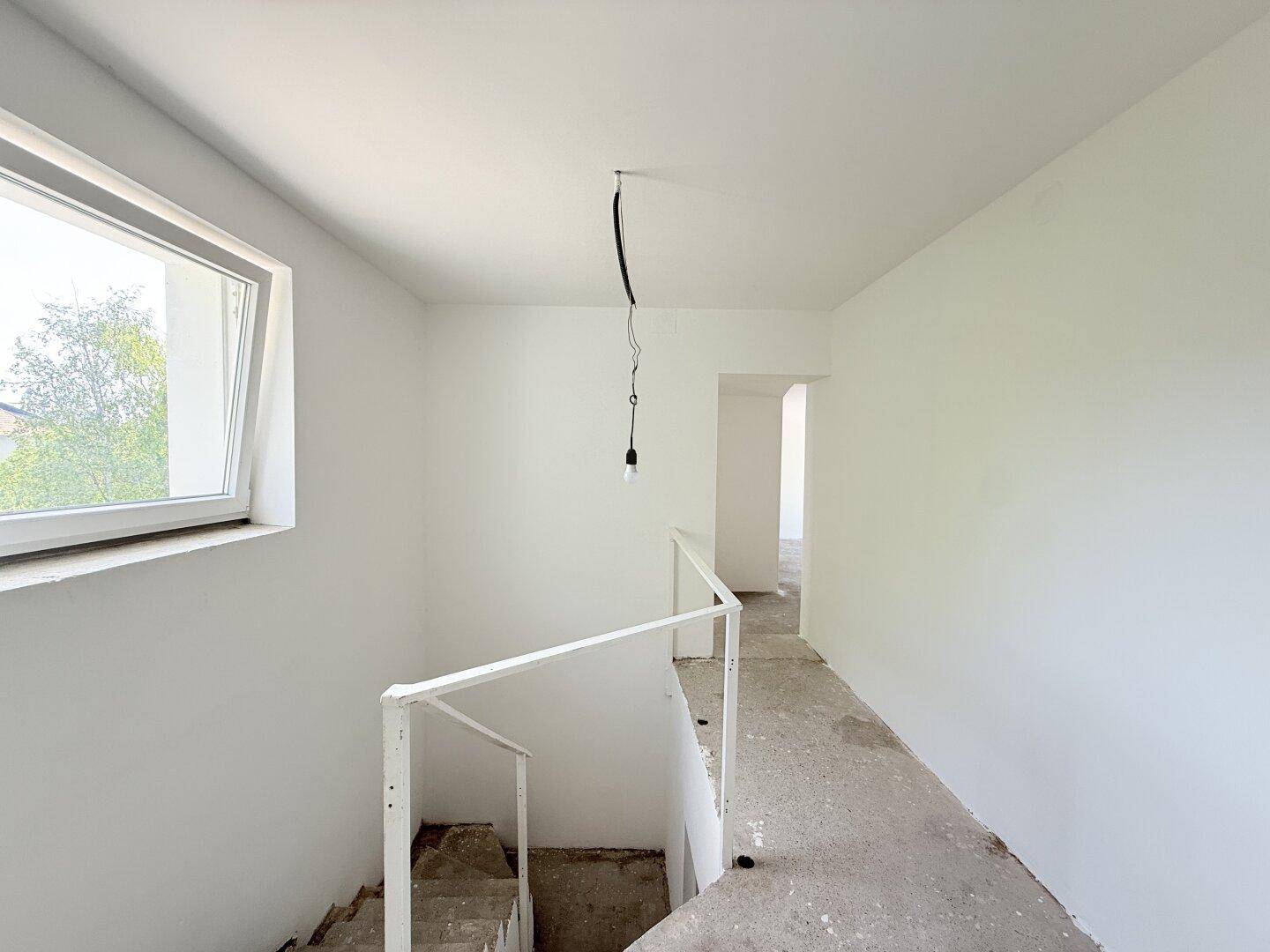 Heller Treppenaufgang mit weiß gestrichenen Wänden und einem Fenster, das Tageslicht hereinlässt.