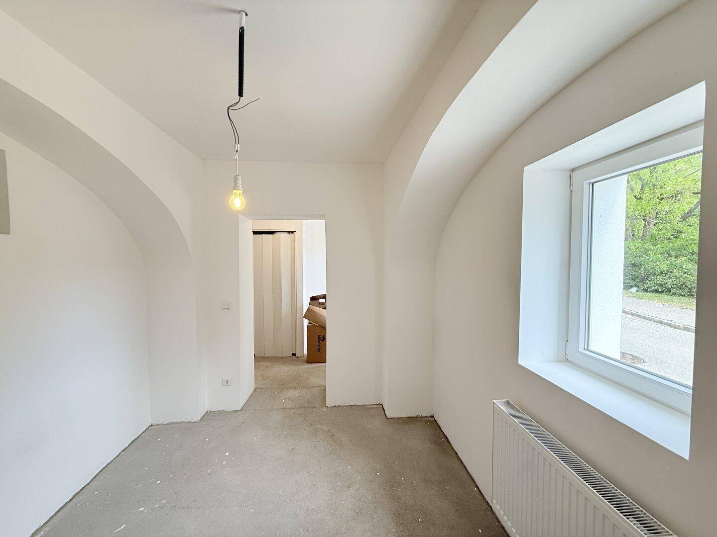 Heller Raum mit Gewölbedecken, Betonboden, großem Fenster und Heizkörper.