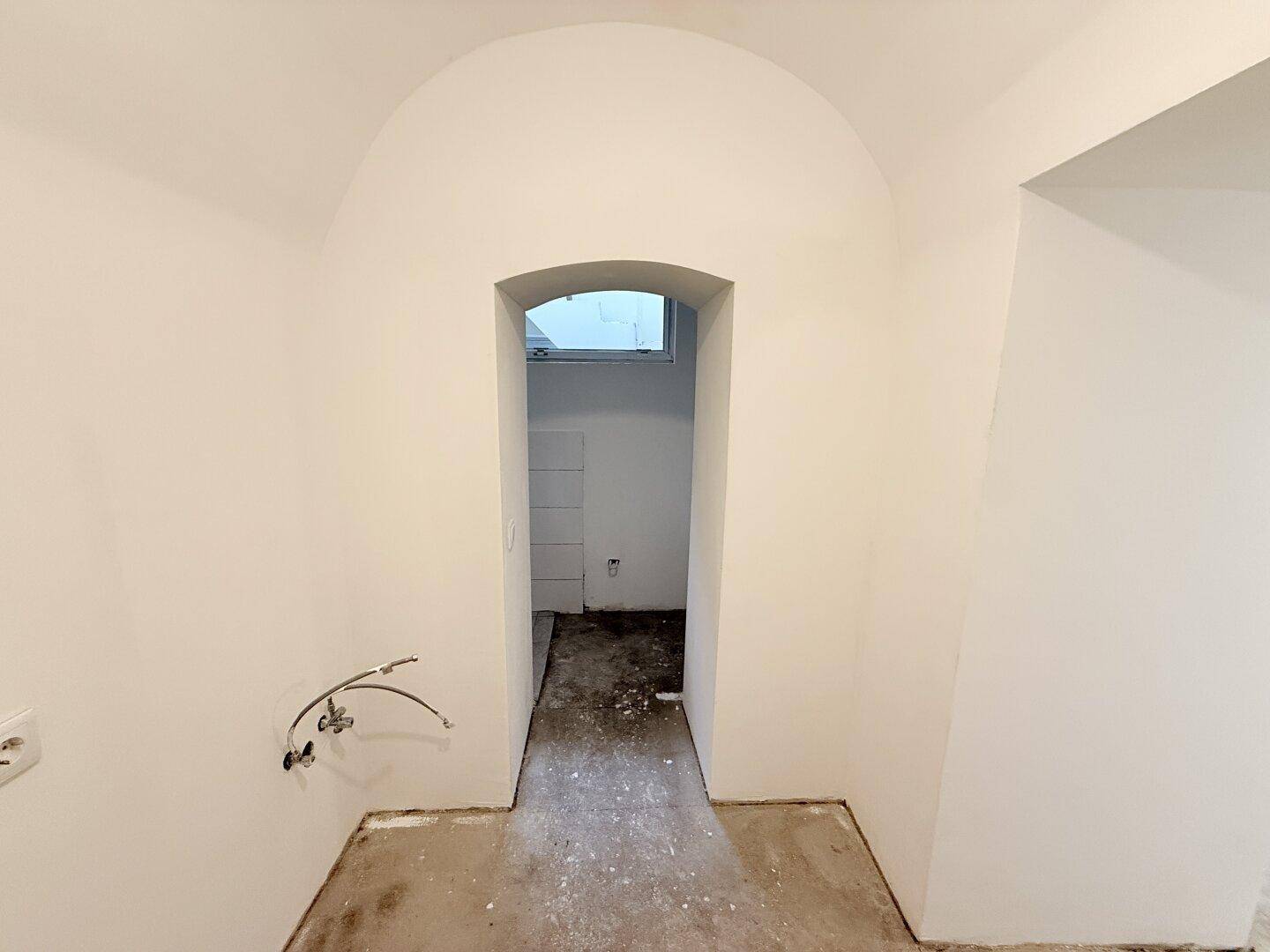 Leerer Raum mit Betonboden, gewölbtem Durchgang und kleinem Fenster, wartet auf Fertigstellung.