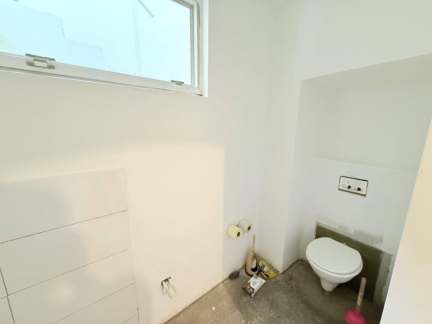 Unfertiges WC mit wandhängender Toilette, Fenster und teilweise gefliesten Wänden.