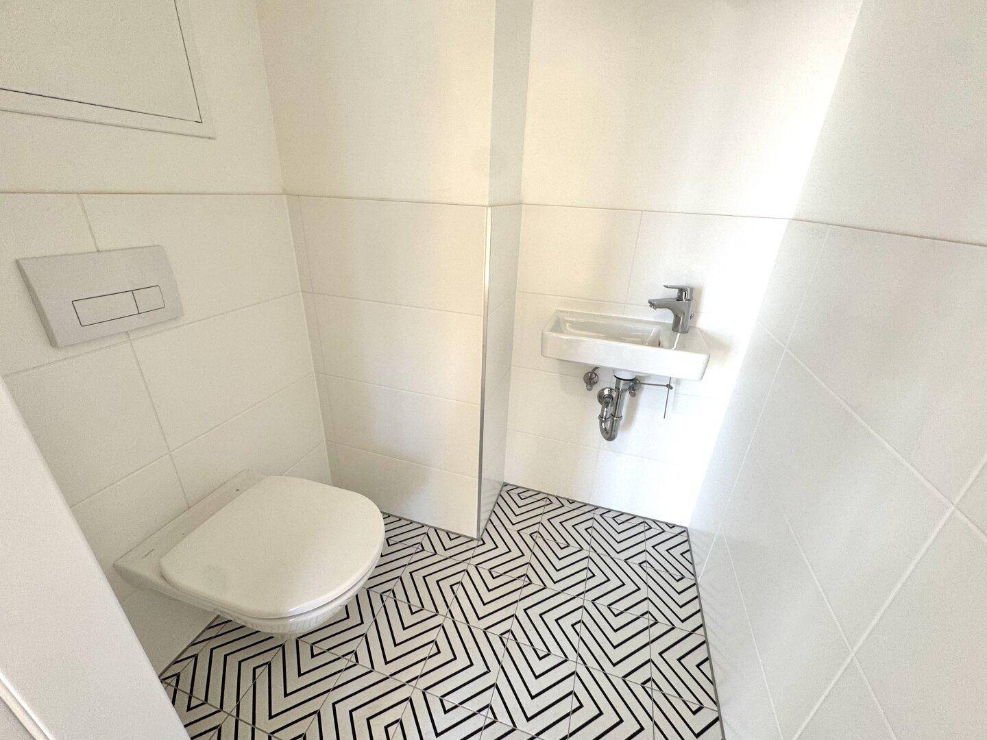 Separates WC mit wandhängender Toilette, kleinem Waschbecken und geometrischen Bodenfliesen.