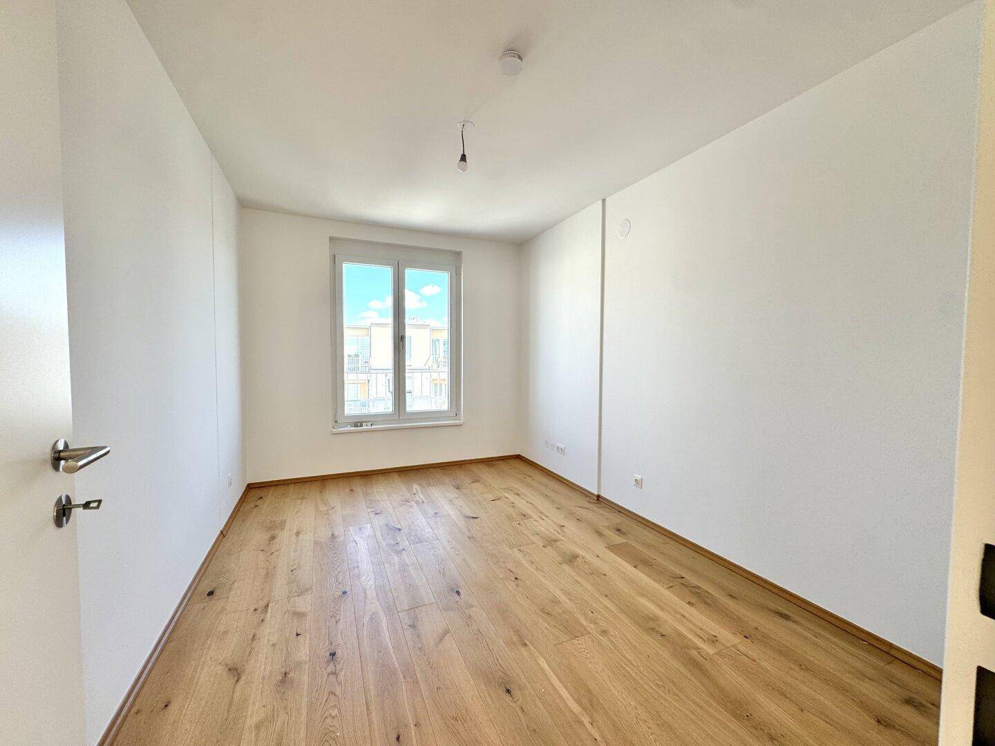 Helles Schlafzimmer mit Holzboden und einem Fenster mit Blick auf die Nachbargebäude.