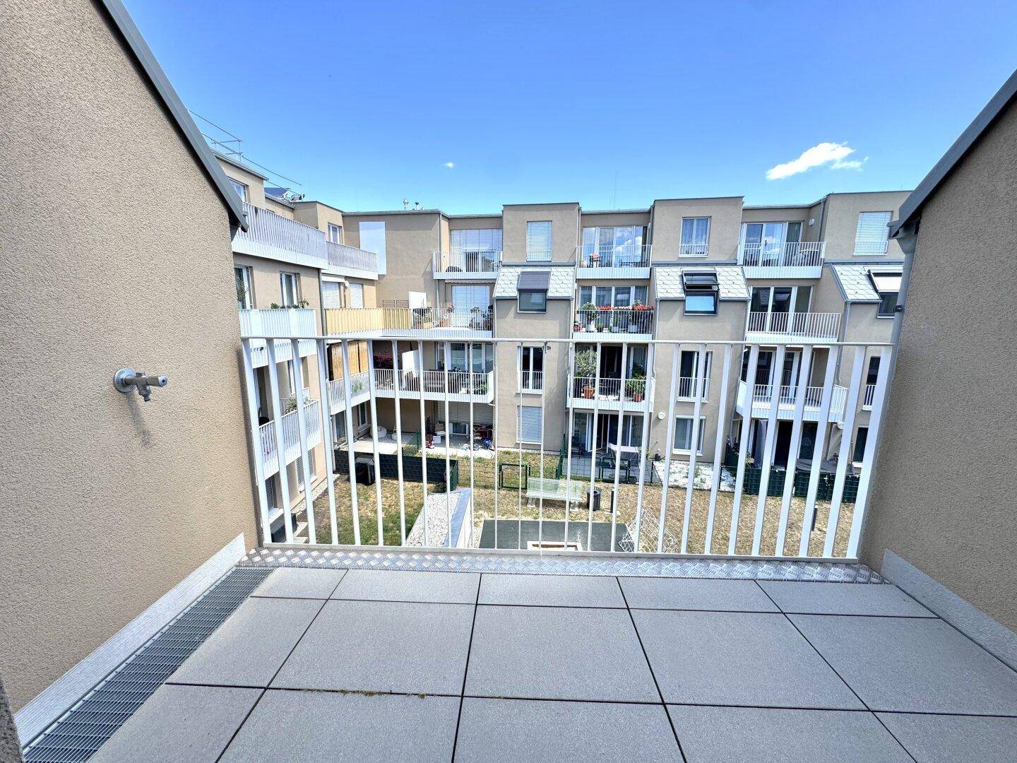 Balkon mit Fliesenboden und Blick in einen begrünten Innenhof, umgeben von Wohngebäuden.