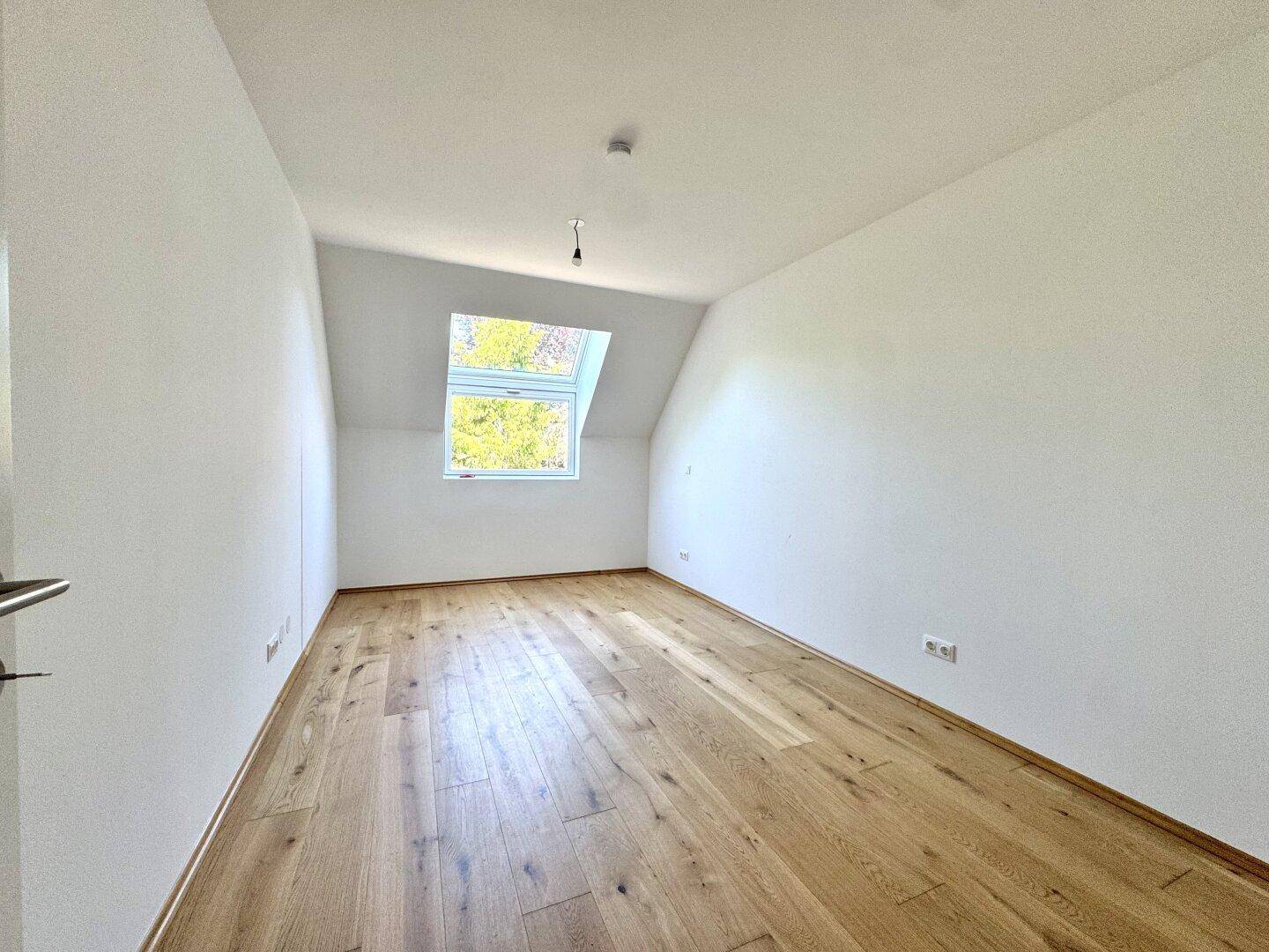 Helles Zimmer im Dachgeschoss mit Holzboden, weiß gestrichenen Wänden und einem Dachfenster.