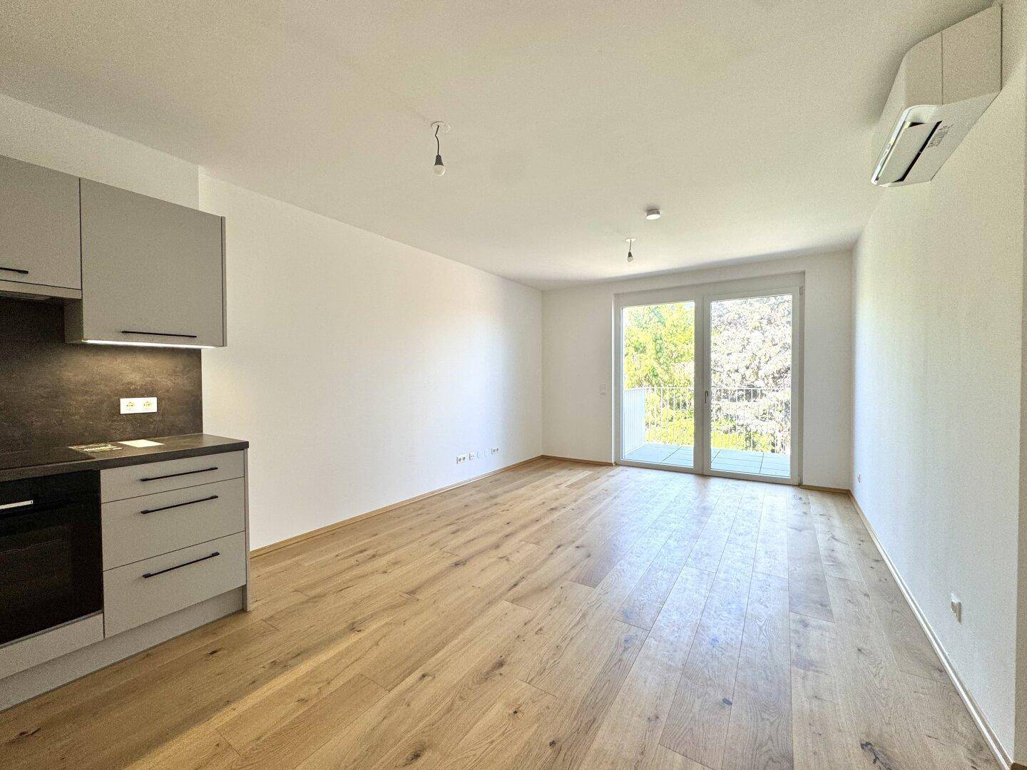 Heller Wohnbereich mit Holzboden, kleiner Küchenzeile und Zugang zu einem Balkon.