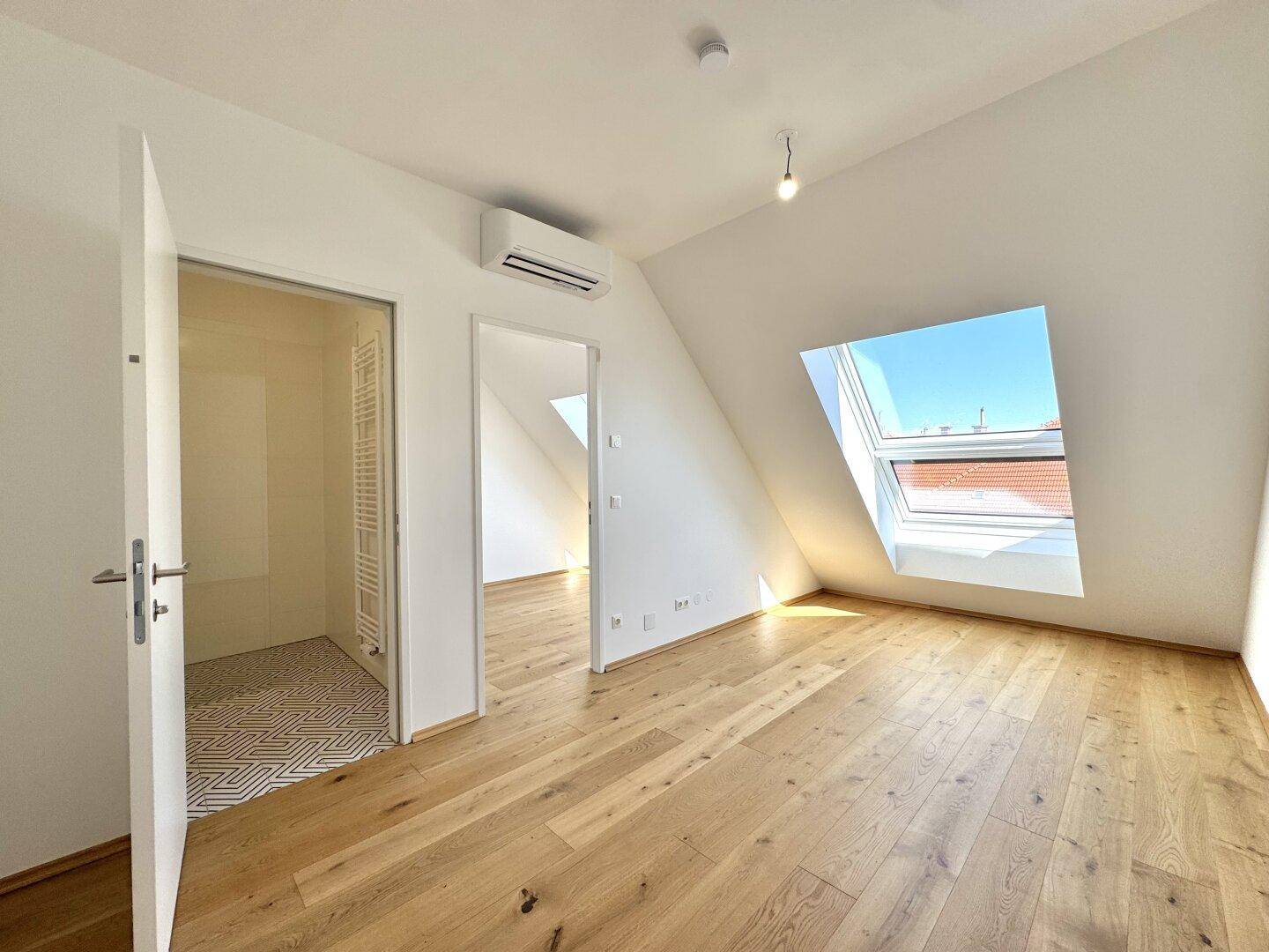 Helles Zimmer mit Holzboden, Dachfenster und Zugang zu einem modernen Badezimmer.