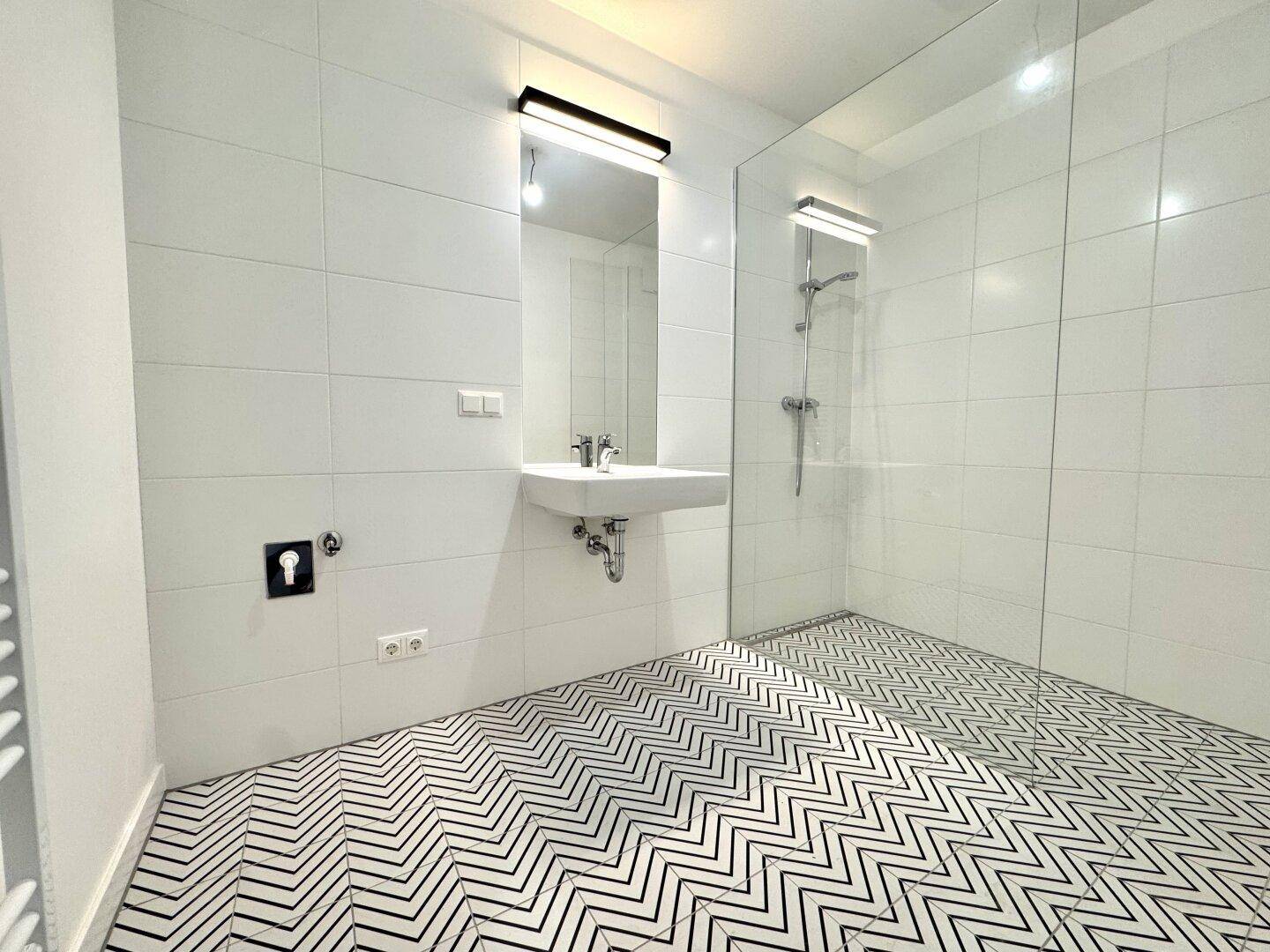 Modernes Badezimmer mit weißen Fliesen, ebenerdiger Dusche und einem stilvollen schwarz-weißen Fliesenboden.