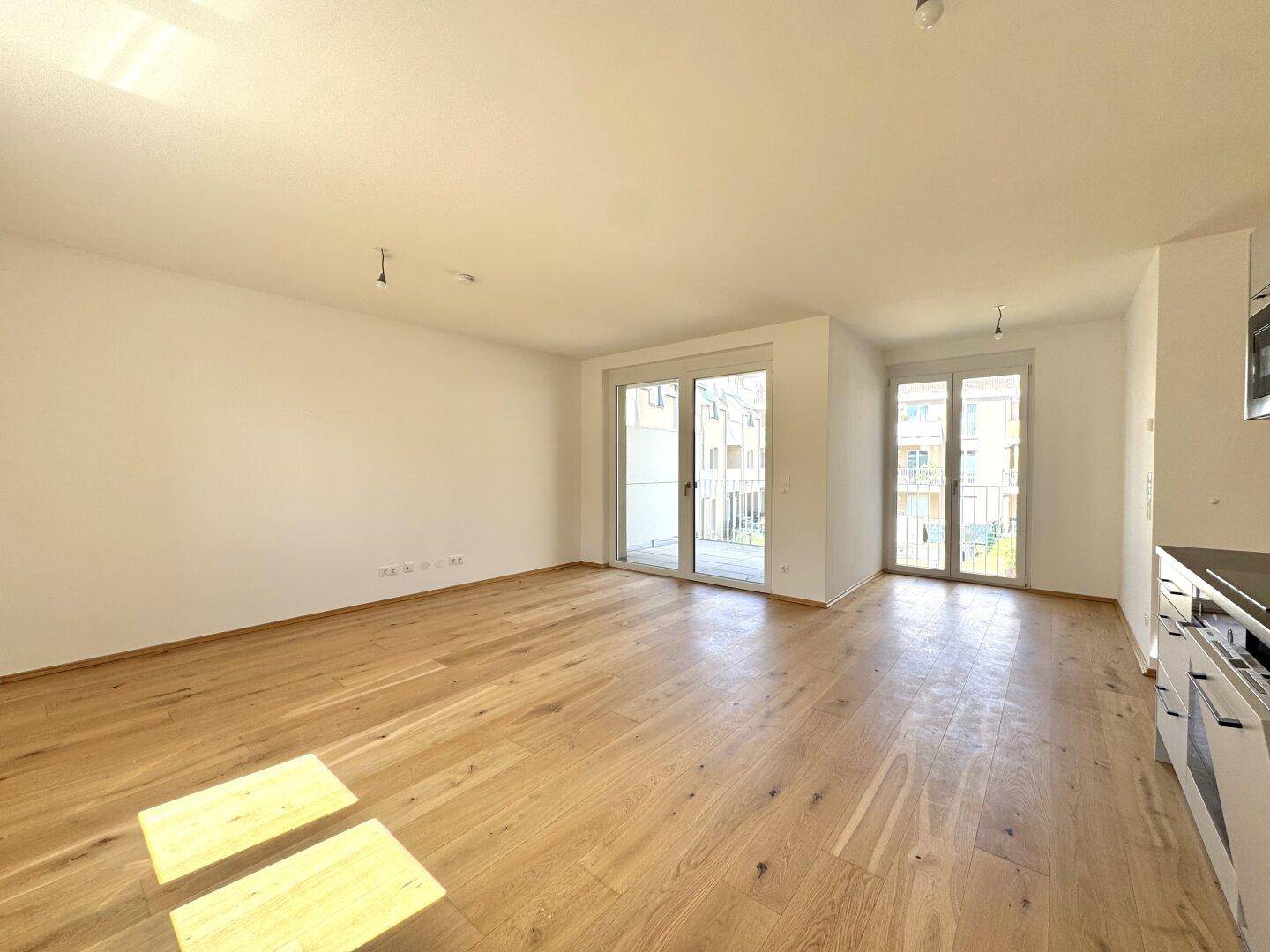 Lichtdurchfluteter Wohnbereich mit Holzboden und großen Fenstern, die auf einen Balkon führen.