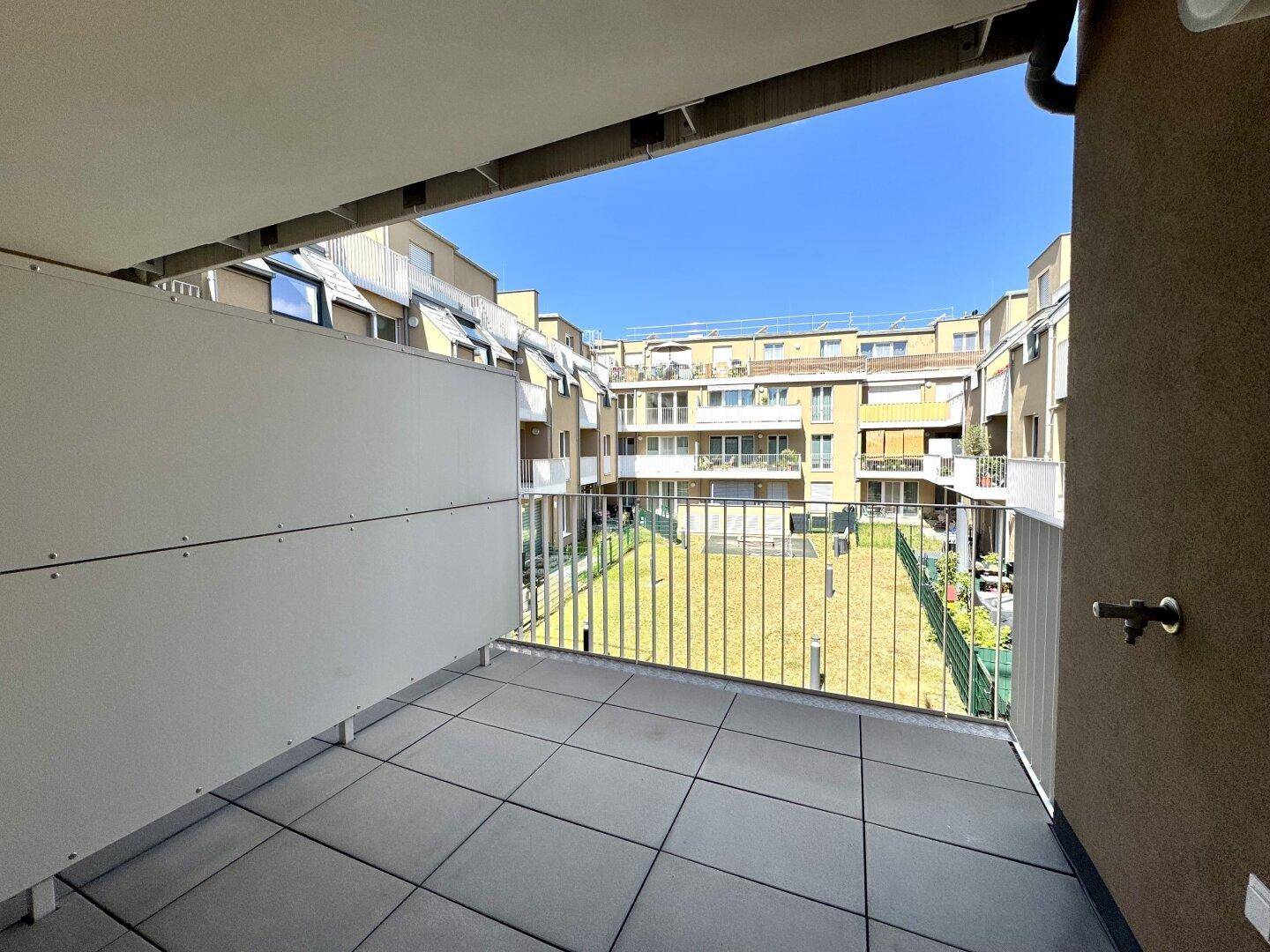 Heller Balkon mit Fliesenboden und Blick auf den grünen Innenhof der Wohnanlage.