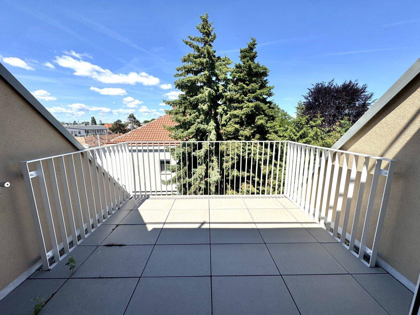 Heller Balkon mit Fliesenboden und weißem Geländer, bietet einen Ausblick auf Bäume und Dächer.