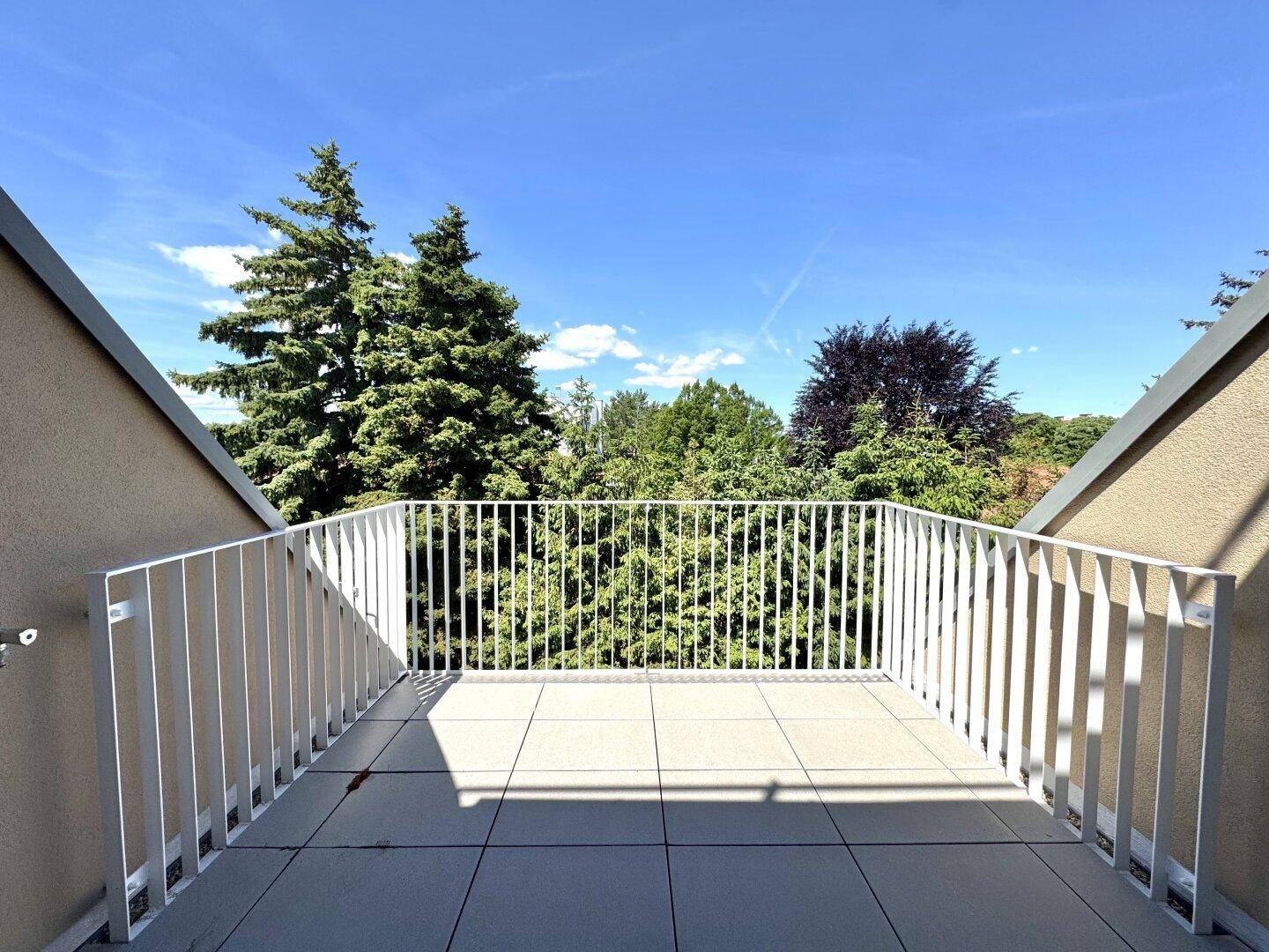 Sonniger Balkon mit Fliesenboden und Blick ins Grüne, umgeben von Bäumen.