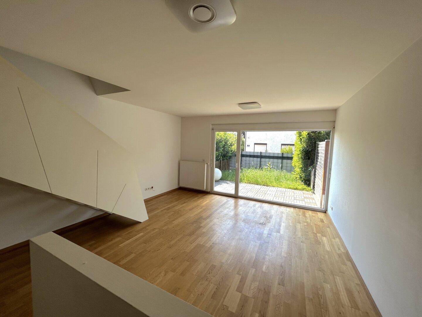 Heller Wohnbereich mit Holzboden und großer Glasschiebetür zum Garten, daneben eine Treppe.