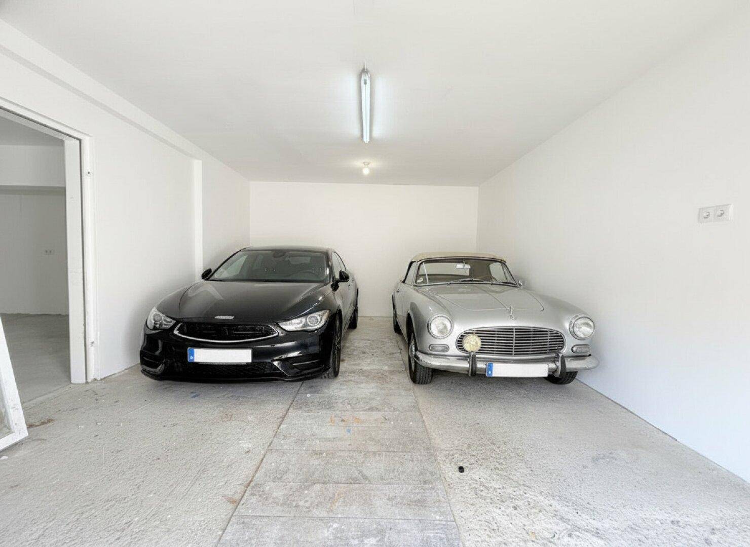 Geräumige Garage mit zwei geparkten Autos, darunter ein Oldtimer, und Betonboden.