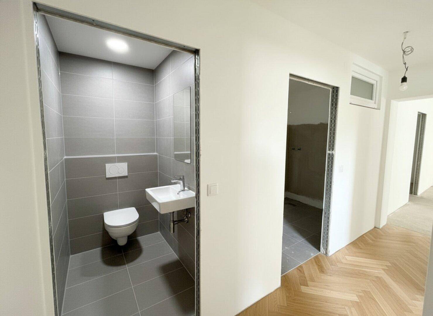 Modernes Gäste-WC mit grauen Fliesen, wandhängendem WC und kleinem Waschbecken.