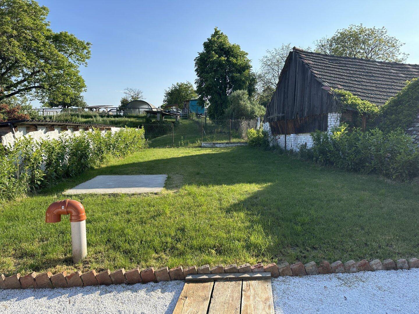 Grüner Gartenbereich mit Rasenfläche und altem Holzschuppen im Hintergrund unter blauem Himmel.