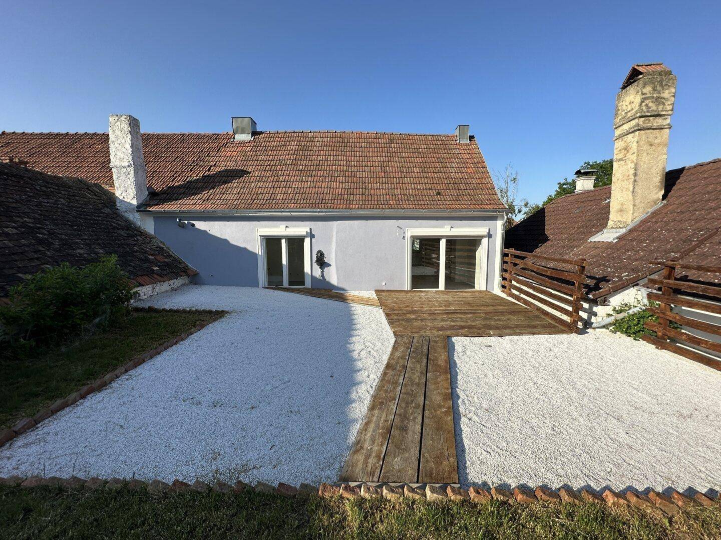 Große Terrasse mit Holzdeck und Kiesfläche hinter einem renovierten Landhaus unter blauem Himmel.