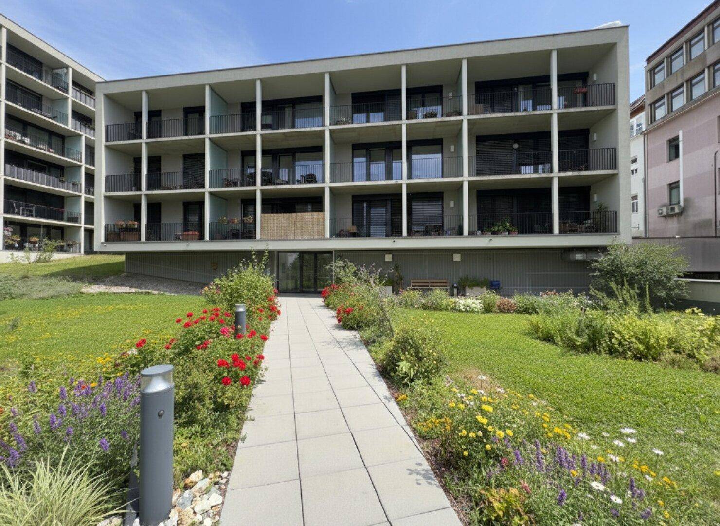 Moderne Wohnanlage mit mehreren Balkonen und einem gepflegten Vorgarten mit Blumenbeeten.