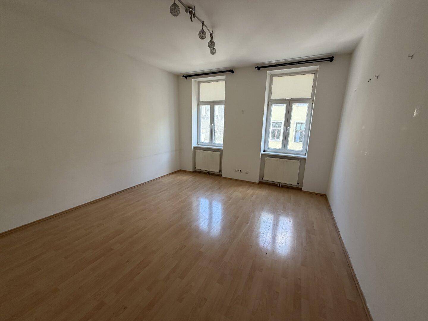 Helles, leeres Zimmer mit Laminatboden und zwei Fenstern, bereit zur individuellen Gestaltung.