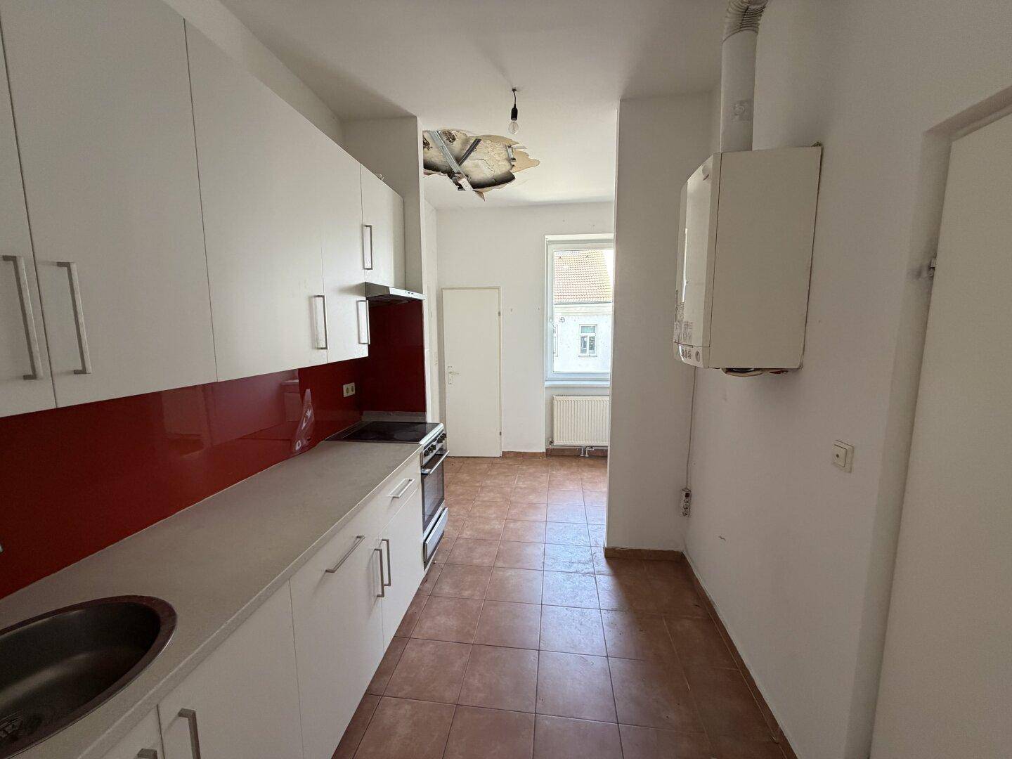 Schmale Küche mit weißen Schränken, roter Rückwand und Gastherme an der Wand.