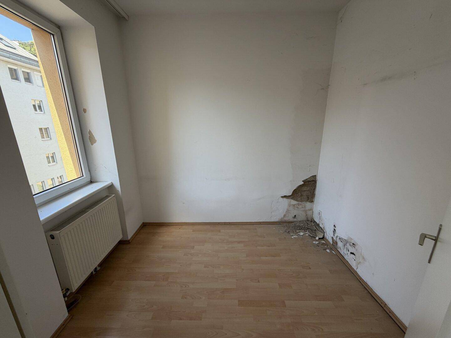 Leeres Zimmer mit Laminatboden und beschädigter Wand, bietet Potenzial zur Renovierung.