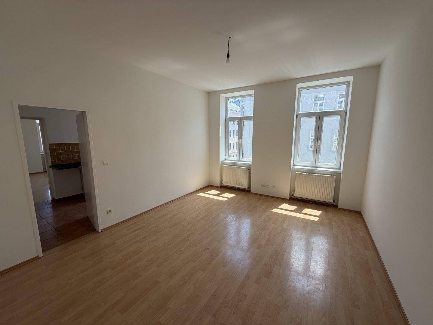 Heller, leerer Raum mit hellem Laminatboden, weißen Wänden und zwei großen Fenstern.