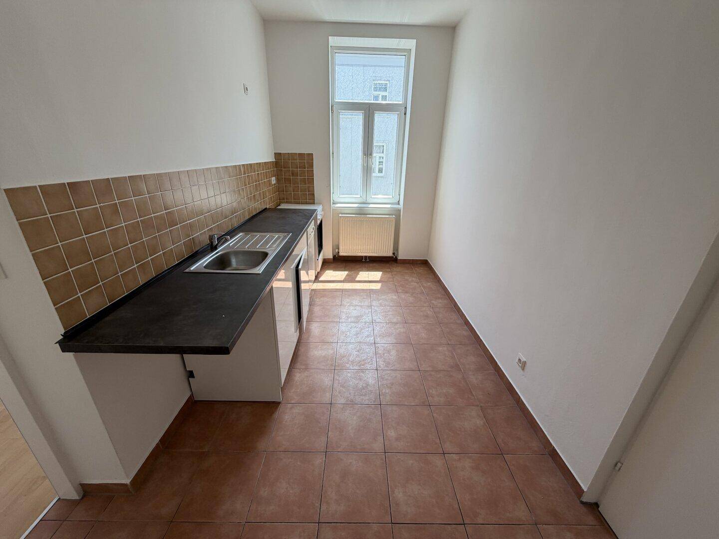 Schmale Küche mit einfacher Küchenzeile, Spüle, braunen Fliesen und einem Fenster.