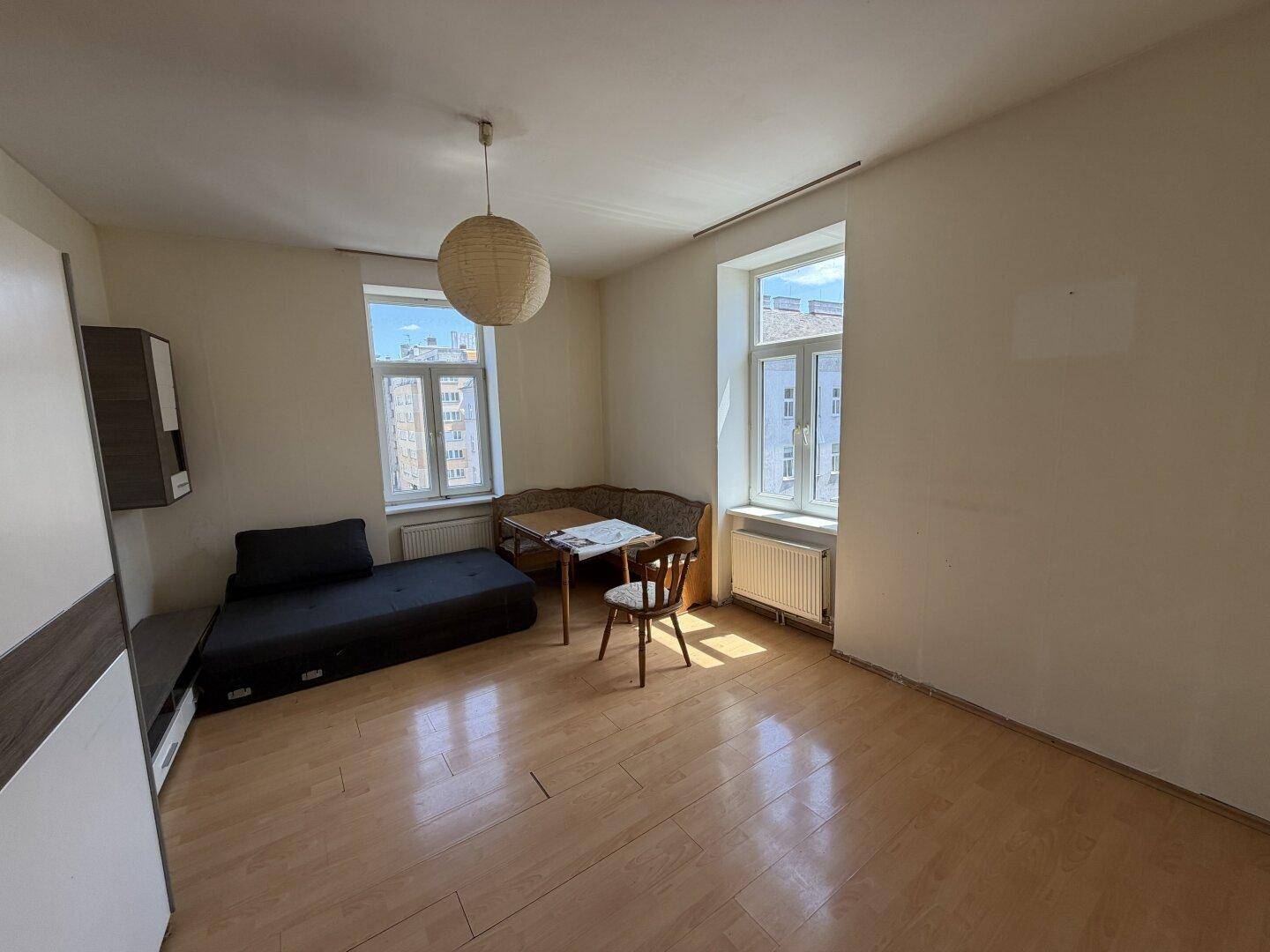 Helles Wohnzimmer mit Holzboden, Schlafsofa und einem kleinen Essbereich am Fenster.
