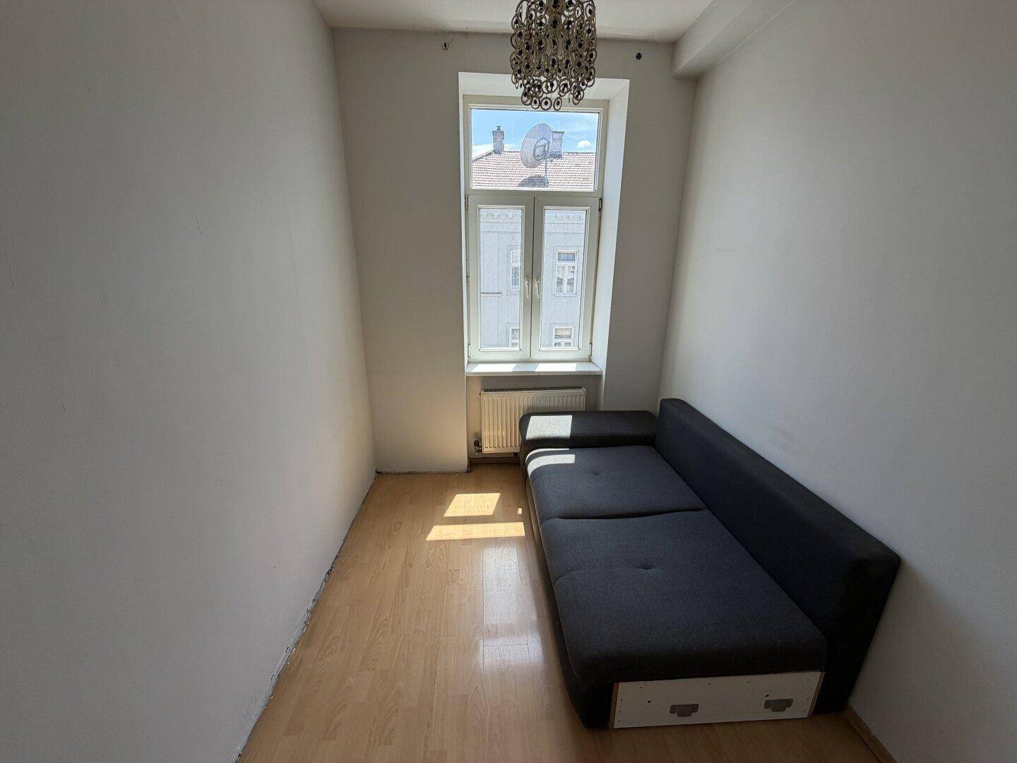 Schmales Zimmer mit Schlafsofa und Holzboden, gut beleuchtet durch ein großes Fenster.