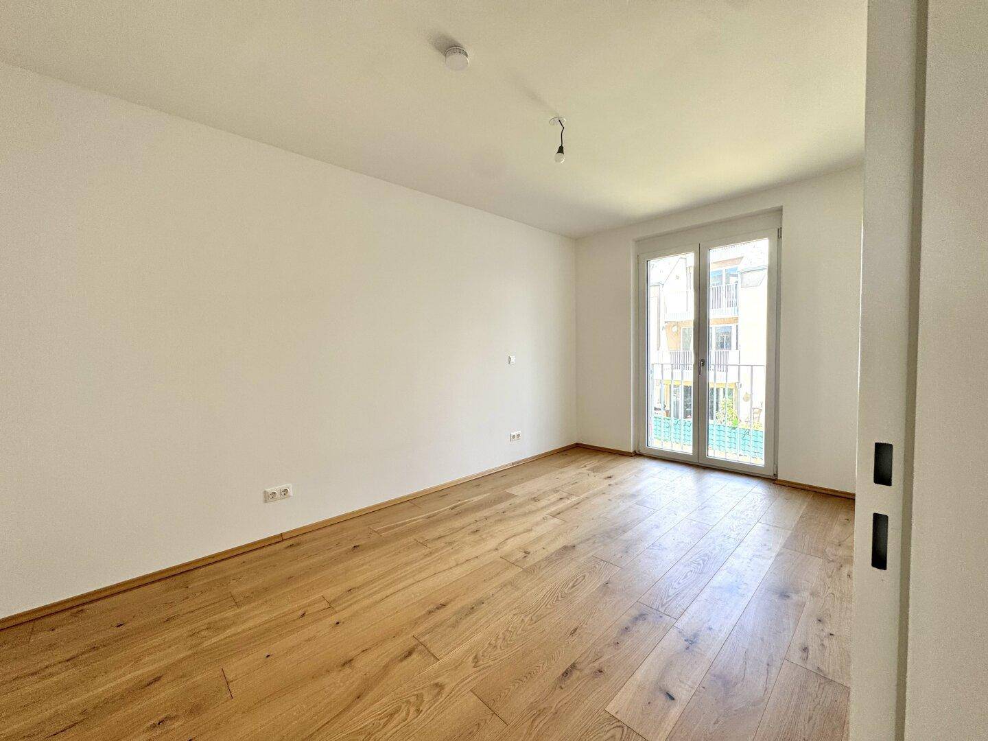 Helles Schlafzimmer mit Holzboden und großem Fenster, das Zugang zum Balkon bietet.