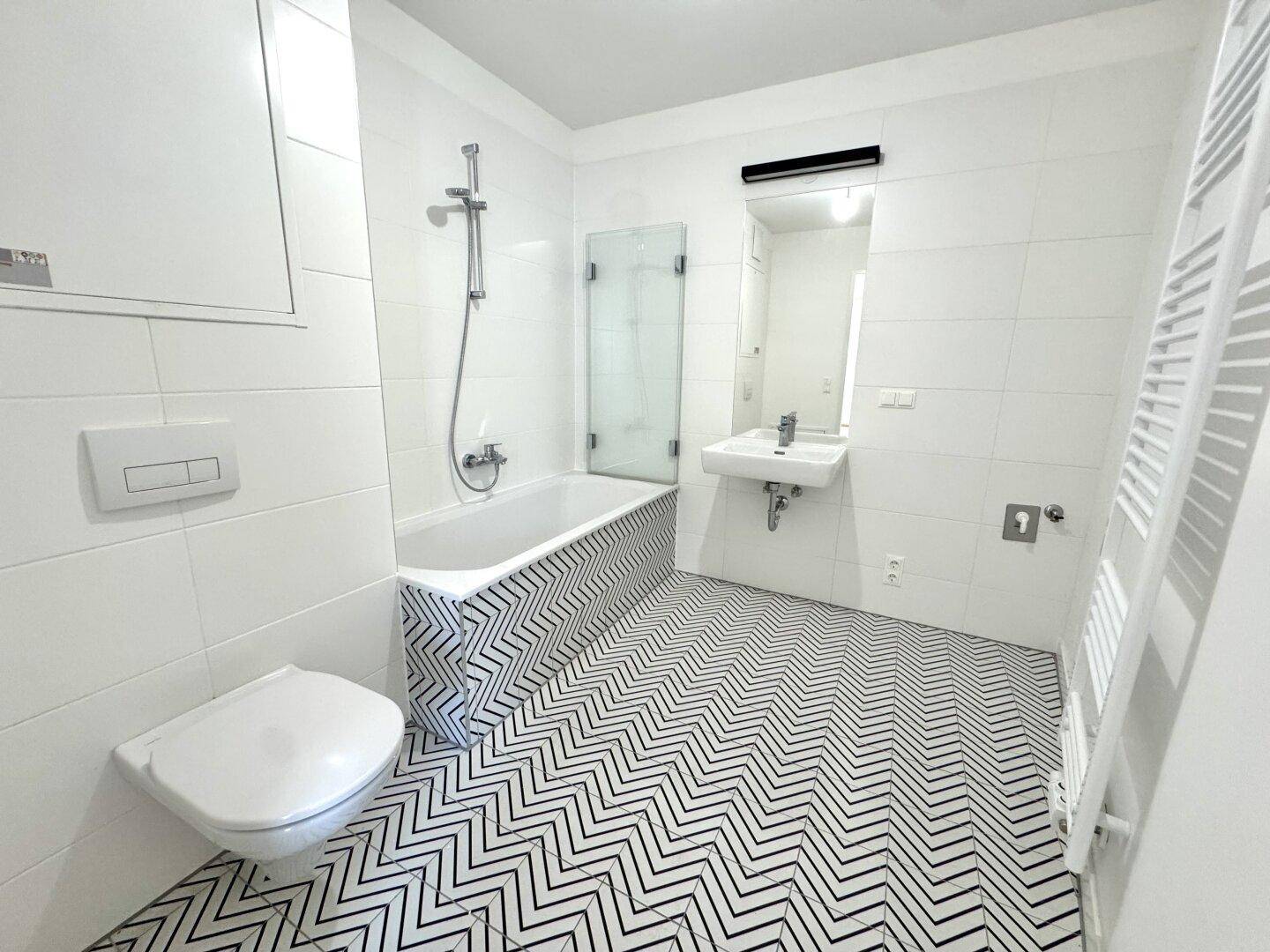Modernes Badezimmer mit weißen Wandfliesen, markantem Bodenmuster und Badewanne mit Duschfunktion.