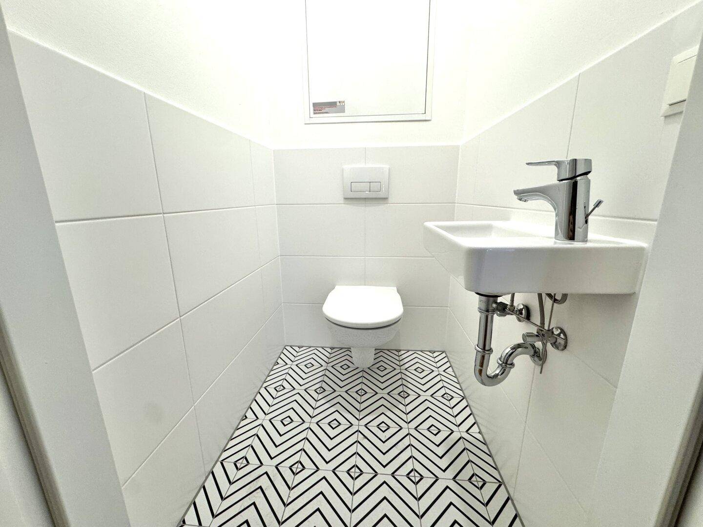 Separates WC mit wandhängender Toilette, kleinem Waschbecken und gemusterten Bodenfliesen.