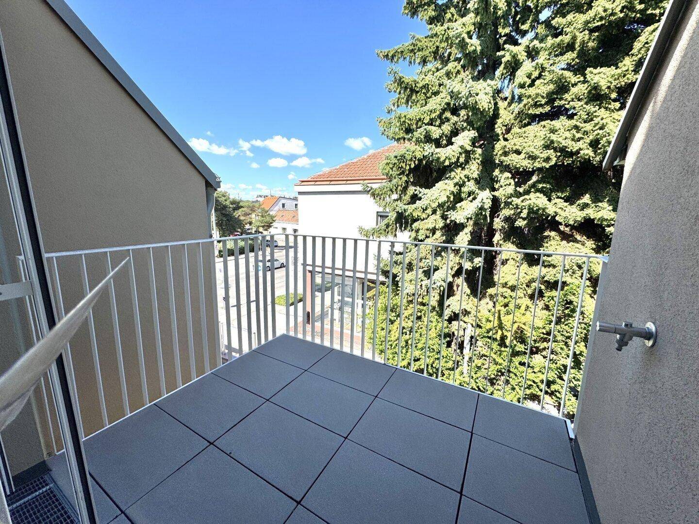 Kleiner Balkon mit grauen Fliesen und weißem Geländer, umgeben von grünen Bäumen.