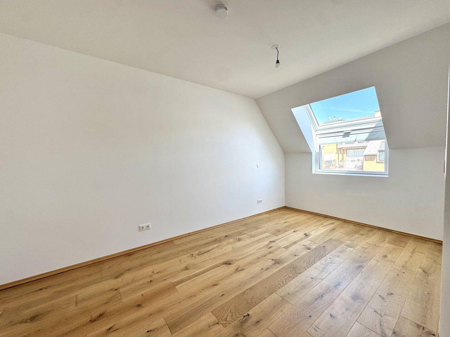 Helles Zimmer im Dachgeschoss mit Holzboden, weiß gestrichenen Wänden und großem Dachfenster.