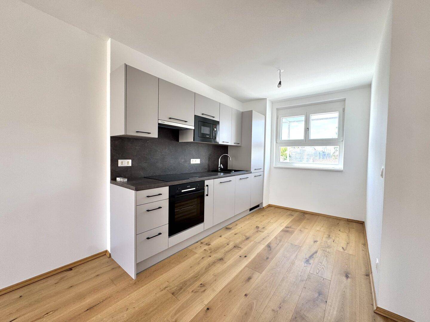 Moderne Küchenzeile mit grauen Schränken, dunkler Arbeitsplatte, integrierten Geräten und Fenster.