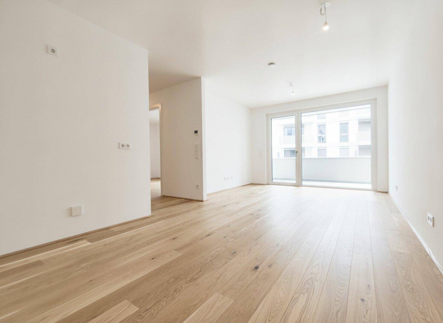 Helles, leeres Wohnzimmer mit neuem Holzparkettboden und Zugang zum Balkon.