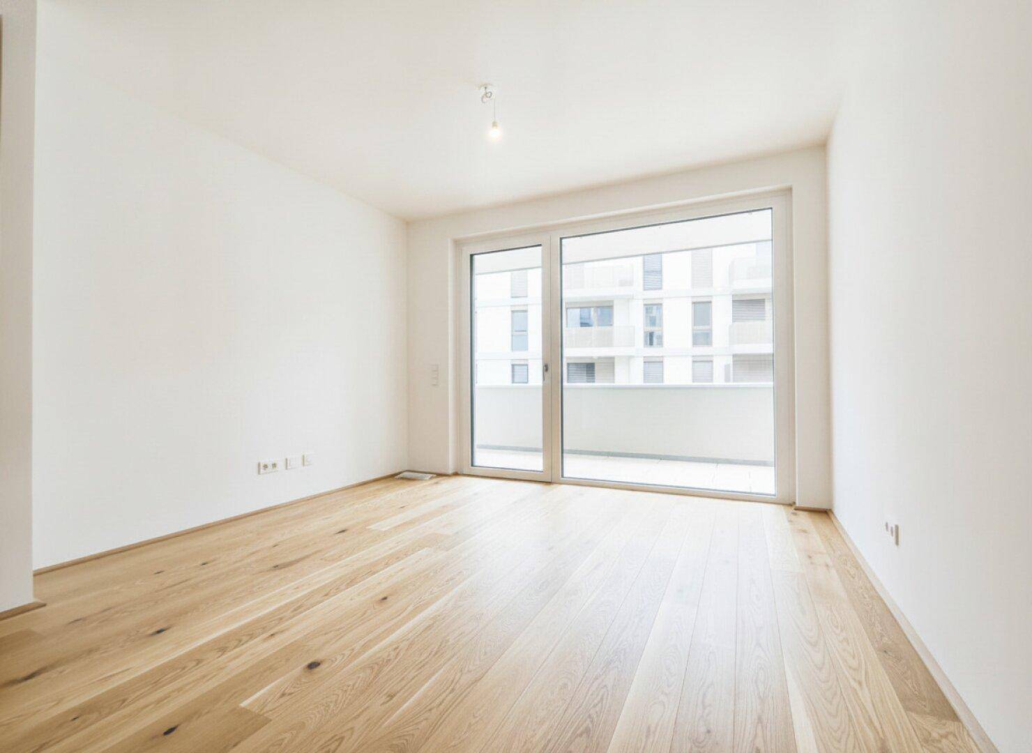 Lichtdurchflutetes Schlafzimmer mit hellem Parkettboden und großem Fenster zum Balkon.